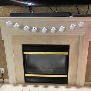 May include: A string of nine white ghost ornaments hanging above a fireplace. The ghosts have black eyes and mouths.