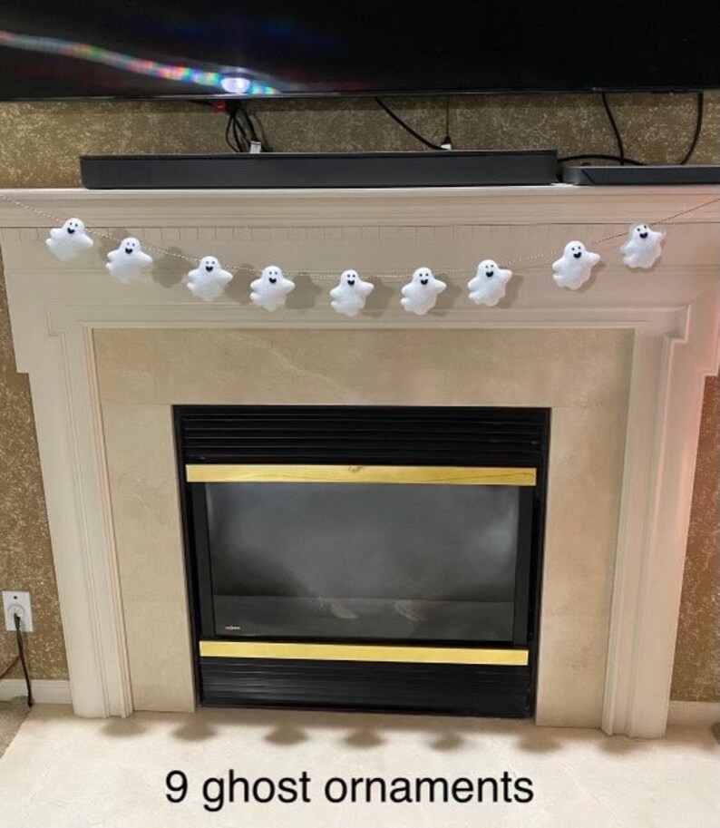 May include: A string of nine white ghost ornaments hanging above a fireplace. The ghosts have black eyes and mouths.