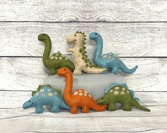 May include: Five felt dinosaur toys in various colors, including green, blue, orange, and beige. The dinosaurs are arranged on a gray surface with a white wood background.