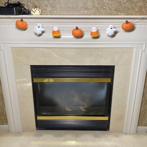 May include: A fireplace mantel decorated with a Halloween garland featuring felt pumpkins, candy corn, and ghosts. The garland is strung with twine and hangs above a black fireplace.