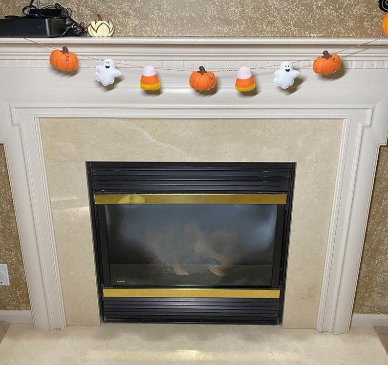 May include: A fireplace mantel decorated with a Halloween garland featuring felt pumpkins, candy corn, and ghosts. The garland is strung with twine and hangs above a black fireplace.