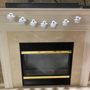 May include: A white felt ghost garland with seven ghosts hanging from a string. The garland is hanging above a fireplace.