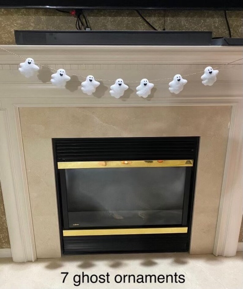 May include: A white felt ghost garland with seven ghosts hanging from a string. The garland is hanging above a fireplace.