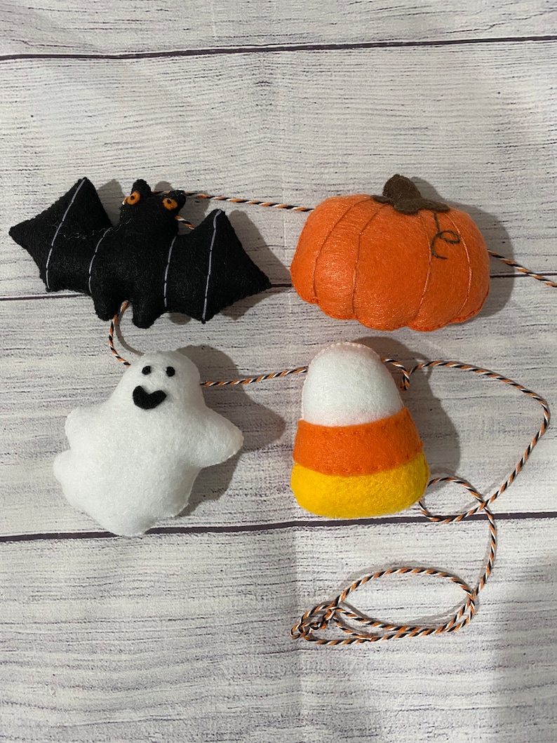 May include: A Halloween garland with felt decorations. The garland features a black bat, an orange pumpkin, a white ghost, and a yellow and orange candy corn.