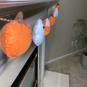May include: A Halloween garland with orange and white felt pumpkins, a ghost, and candy corn hanging from a white mantel.