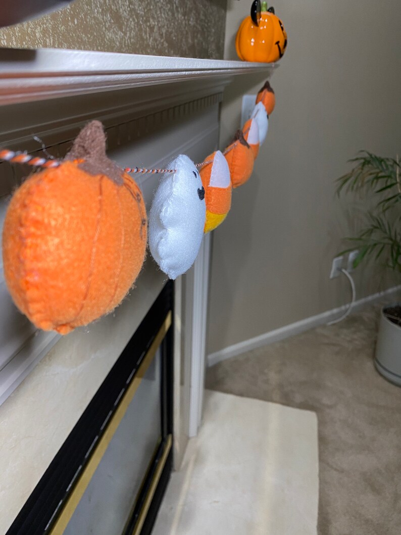 May include: A Halloween garland with orange and white felt pumpkins, a ghost, and candy corn hanging from a white mantel.