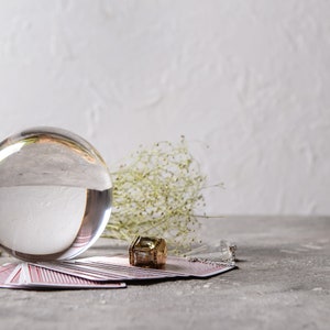 May include: A clear crystal ball sits on a stack of tarot cards with a gold pendant charm. The background is a white wall with a sprig of dried flowers.