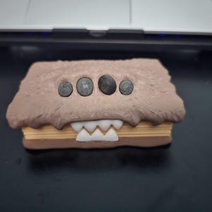 May include: A brown and beige monster-shaped box with five black eyes and a row of white teeth. The box is made of a textured material and has a slightly rounded shape.