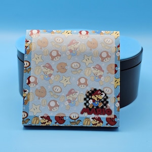 May include: A light blue sticker sheet featuring a repeating pattern of Mario characters, coins, stars, and mushrooms. The word "MARIO" is displayed in a checkered box. The sticker sheet is on a blue background.