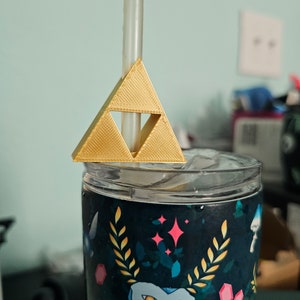 May include: A gold-colored plastic triangle with a straw going through it. The triangle is on top of a clear tumbler with a blue and green owl design.