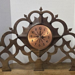 May include: A wooden mantel clock with a copper-colored clock face. The clock has a decorative, openwork wooden frame with an ornate, symmetrical design. The clock face features black numerals and hands.
