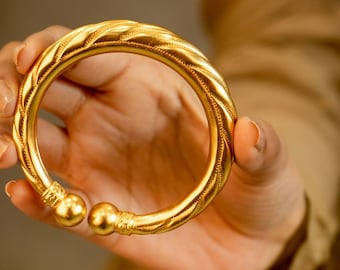 May include: A gold-colored bangle bracelet with a textured, braided design. The bracelet has a rounded shape with decorative detailing and two spherical end caps. The jewelry is held in a hand.