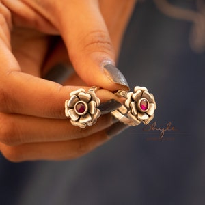 May include: Two silver toe rings with a flower design and a pink gemstone in the centre. The rings are held in a hand.