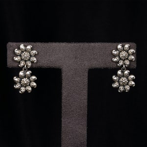 May include: A pair of silver dangle earrings with a floral design. The earrings are on a gray display stand.