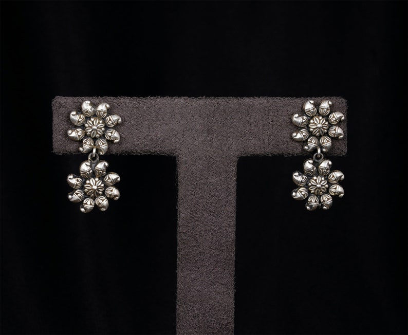 May include: A pair of silver dangle earrings with a floral design. The earrings are on a gray display stand.