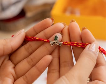 May include: A red and yellow thread bracelet with a silver charm featuring the word "Raksha" in Hindi script and a green stone.