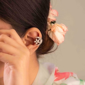 Op de afbeelding: Zilverkleurige oormanchet met een bloemmotief. De manchet heeft de vorm van een halve maan met een bloem in het midden, versierd met kleine, heldere steentjes. De sieraden worden op het oor gedragen en tonen de delicate details en elegante stijl.