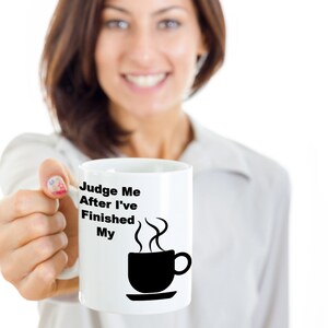 Op de afbeelding: Witte keramische koffiemok met zwarte tekst die "Judge Me After I've Finished My" luidt en een zwart silhouet van een koffiekopje met stoom die eruit opstijgt.