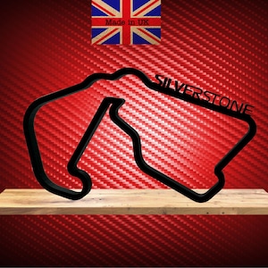Puede incluir: Silueta negra de metal de la pista de carreras de Silverstone, montada en un estante de madera sobre un fondo rojo de fibra de carbono. El texto "SILVERSTONE" se muestra en la pista.