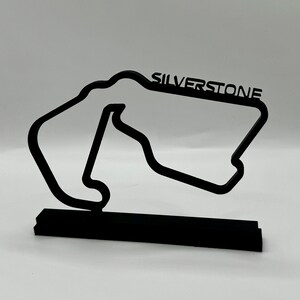 May include: Black metal model of the Silverstone racetrack with the name "SILVERSTONE" printed on the track. The model is on a black base.