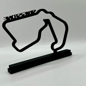 May include: Black 3D printed model of the Monza race track with the word "MONZA" printed on the top. The model is on a black base.