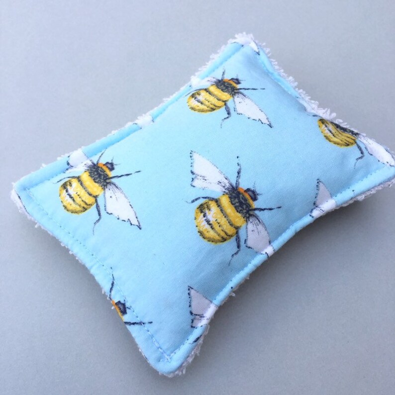 May include: A light blue rectangular sponge with a repeating pattern of yellow and black bees with white wings. The sponge has a white, fringed border.