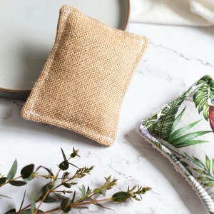 May include: Two rectangular dish scrubbers. One is made of tan burlap with white stitching. The other has a white background with a green tropical leaf and bird pattern. Both are on a white marble surface.