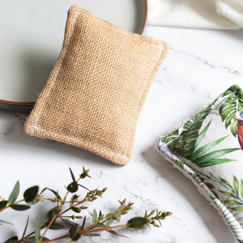 May include: Two rectangular dish scrubbers. One is made of tan burlap with white stitching. The other has a white background with a green tropical leaf and bird pattern. Both are on a white marble surface.