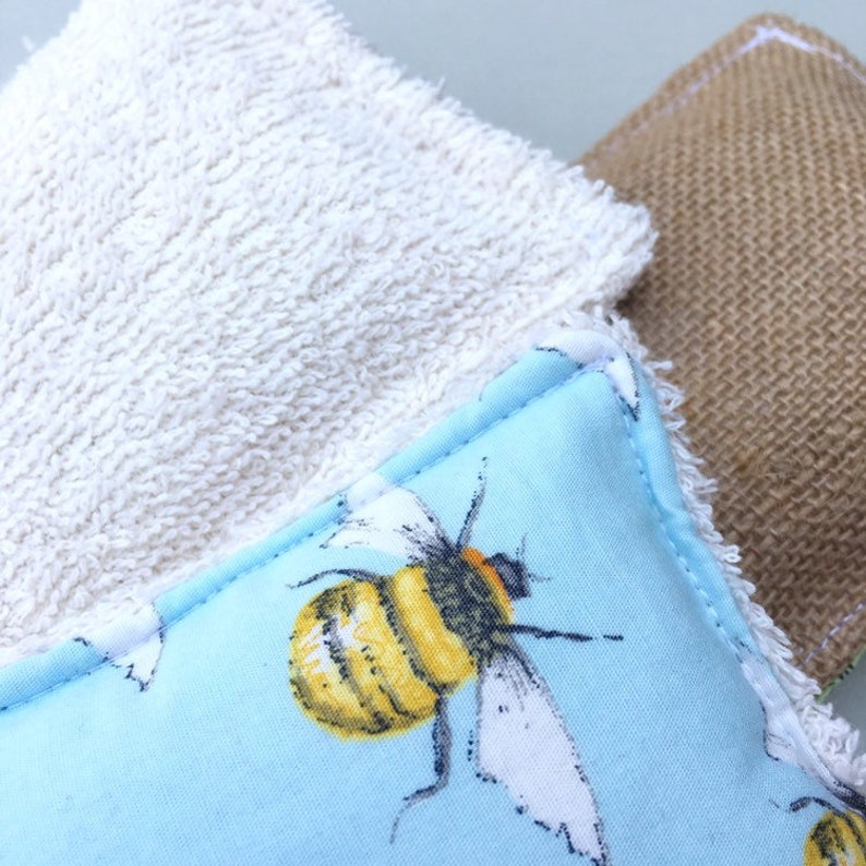 May include: Close-up of three reusable cleaning pads. One is white terry cloth, another is light blue with a yellow and black bee print, and the third is a brown burlap material. The pads are square-shaped and have a fringed edge.