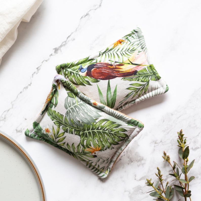 May include: Two square, decorative dish scrubbers with a tropical leaf and bird print. The scrubbers feature a white background with green leaves, orange flowers, and a brown and blue bird. The scrubbers are on a white marble surface.