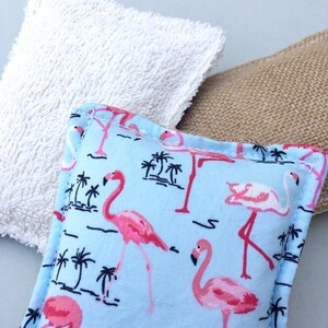 May include: Close-up of three square fabric items. The front item is a light blue pillow with a pink flamingo and palm tree pattern. A white terry cloth square and a burlap square are behind the flamingo pillow.