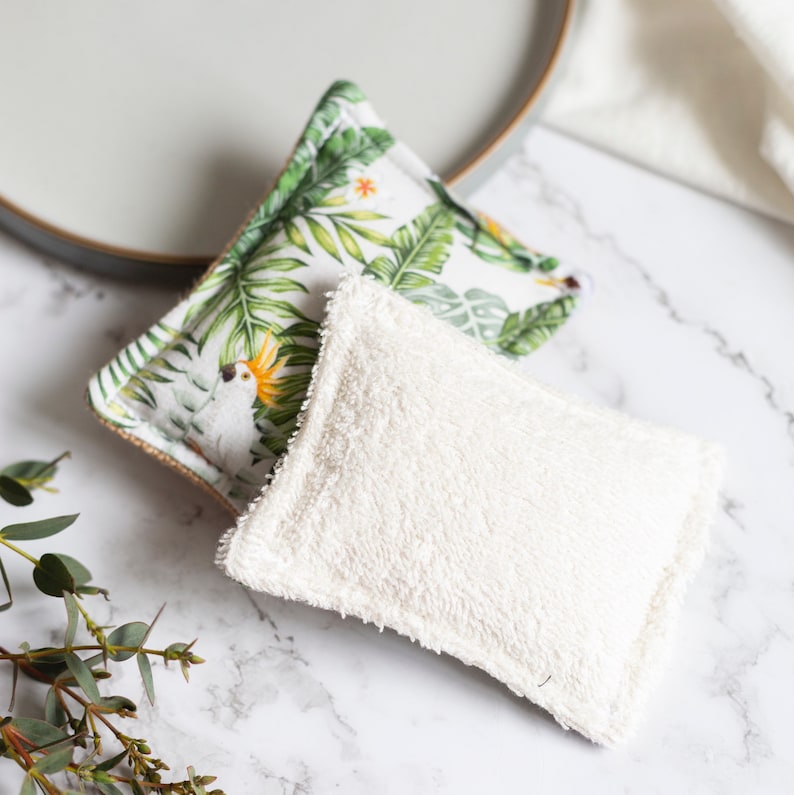 May include: Two square reusable cleaning pads. One pad features a white background with a green tropical leaf and parrot print. The other pad is white and fluffy. The pads are on a marble surface.