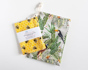 May include: Two waterproof coffee cup pouches. One is yellow with a honeycomb pattern and bee illustrations. The other is white with a tropical leaf and toucan design. Both have a drawstring closure. The yellow pouch has a white label with text.