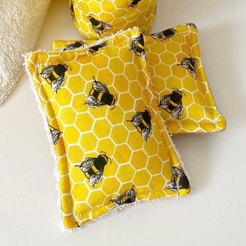 May include: Yellow and white honeycomb pattern fabric with black and yellow bee illustrations. The fabric is used to make two square scrubbies with white trim. A matching round item is in the background.