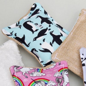 May include: A collection of reusable cleaning pads with various patterns. One pad features a light blue background with black and white penguins. Another has a pink background with unicorns and rainbows. A third pad is made of a natural burlap material.