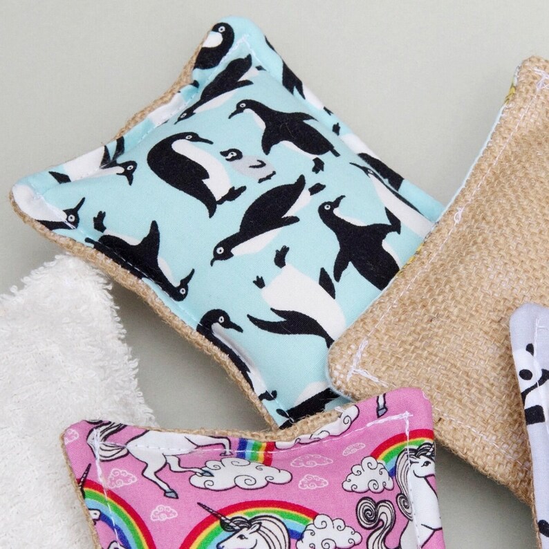 May include: A collection of reusable cleaning pads with various patterns. One pad features a light blue background with black and white penguins. Another has a pink background with unicorns and rainbows. A third pad is made of a natural burlap material.
