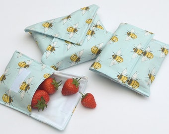 May include: Reusable light blue snack bags with a repeating pattern of yellow and black bees. One bag is open, filled with red strawberries. The bags have a velcro closure. The bags are made of fabric.