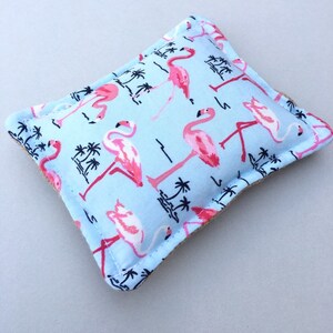 May include: A light blue, rectangular decorative pillow featuring a repeating pattern of pink flamingos and black palm trees. The pillow has a beige border and is likely made of fabric.