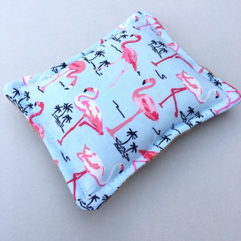 May include: A light blue, rectangular decorative pillow featuring a repeating pattern of pink flamingos and black palm trees. The pillow has a beige border and is likely made of fabric.