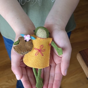 May include: A small handmade felt frog doll featuring a tan face, green limbs, and a yellow tunic with a pink embroidered flower. The frog holds a brown felt basket adorned with a small pink flower and blue leaves, stitched with visible thread.