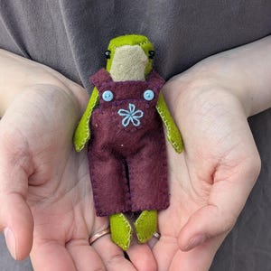 May include: A small handmade felt frog doll with a light green body and black bead eyes. The frog is wearing dark purple felt overalls featuring two light blue buttons at the shoulders and a light blue embroidered flower on the chest.