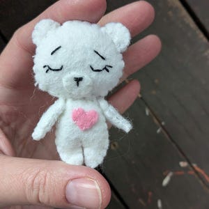 May include: A small, handmade white felt bear with a pink heart on its chest. The bear has closed eyes and a black stitched nose and mouth. The bear is being held in a hand against a dark wooden background.