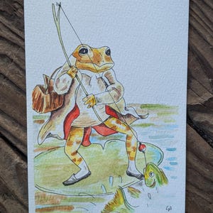 May include: Watercolor illustration of a frog dressed in a coat and vest, fishing with a bamboo rod. The frog stands on a lily pad, with a fish biting the hook. The artwork has a whimsical, storybook style.