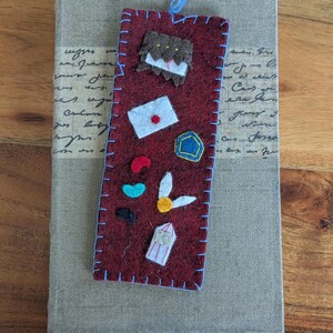 May include: A rectangular dark red felt bookmark with light blue stitched edges and a matching blue thread tassel. It features small felt appliqués including a brown creature, a white envelope with a red seal, a blue pentagon, colorful beans, a golden snitch, and a striped ticket.