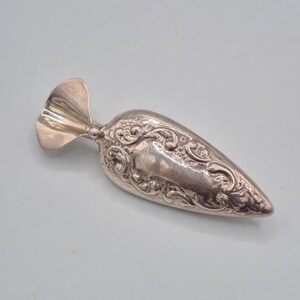 May include: A silver-toned metal sugar scoop featuring an elongated, tapered bowl with intricate embossed scrollwork and floral patterns. The handle is short and wide, shaped like a fan or a shell, connecting to the main body with a small ball.