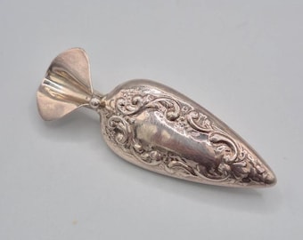May include: A silver-toned metal sugar scoop featuring an elongated, tapered bowl with intricate embossed scrollwork and floral patterns. The handle is short and wide, shaped like a fan or a shell, connecting to the main body with a small ball.