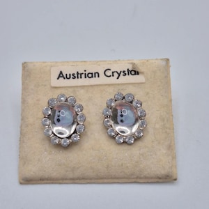 May include: A pair of oval-shaped stud earrings featuring a central clear cabochon stone surrounded by a halo of small, round, clear crystals. The earrings are mounted on a beige display card with the text Austrian Crystal printed on a label.