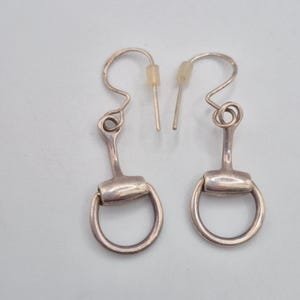 Puede incluir: Un par de pendientes plateados con diseño de bocado de caballo. Cada pendiente tiene un gancho curvo y un pequeño poste cilíndrico. La parte del bocado es un rectángulo redondeado con un anillo circular en cada extremo.