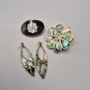 Op de afbeelding: Een collectie sieraden bestaande uit een ovale broche met een zwarte achtergrond en witte bloeminleg, een ronde bloemvormige broche met iriserende abalone schelpsegmenten en een paar druppelvormige oorbellen met abalone schelp inleg.
