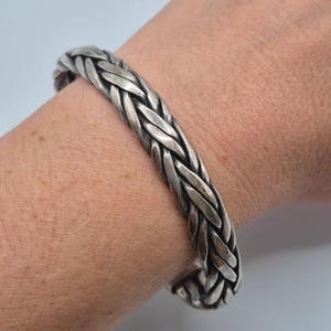 May include: A silver-toned metal bracelet featuring a braided, woven design with a polished finish. The band has a continuous, interlocking pattern that wraps around the wrist, showcasing a textured, multi-strand appearance throughout the piece.